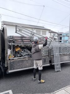 三木市の外壁塗装現場で足場資材をトラックから運ぶ職人の背中
