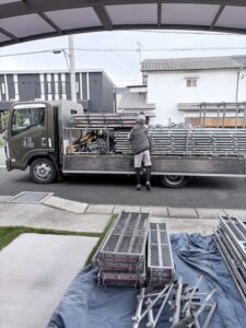 三木市の外壁塗装現場で足場資材をトラックから運ぶ職人の背中