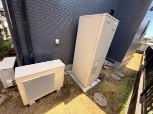 "施工後のエコキュート設置場所の様子。ニンバスに塗り替えられた外壁の前に、新しく設置されたダイキン製エコキュートとヒートポンプユニットが美しく収まっている。"