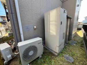 施工前のエコキュート設置場所の様子。グレーのサイディング外壁の前に旧型のエコキュートとヒートポンプユニットが設置されている。"